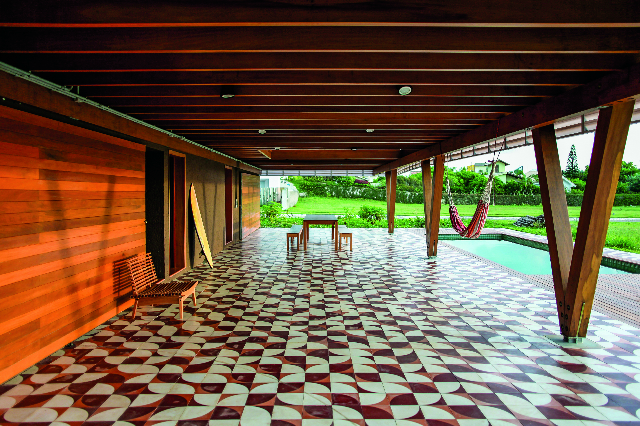 Esta casa de veraneio, projetada pelo Una Arquitetos em Florianópolis, é suspensa, criando uma ampla varanda. Nela, o piso de ladrilho hidráulico forma um grande mosaico que se estende para o exterior, contornando a piscina 