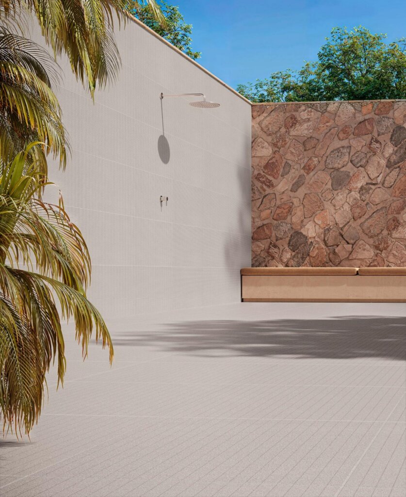 rea externa minimalista e moderna sob céu azul, com chuveiro de parede cromado fixado em parede texturizada clara. À direita, uma parede de pedra rústica irregular com um banco de alvenaria e assento estofado bege. No primeiro plano, piso de área externa texturizado com sombras de árvores projetadas.