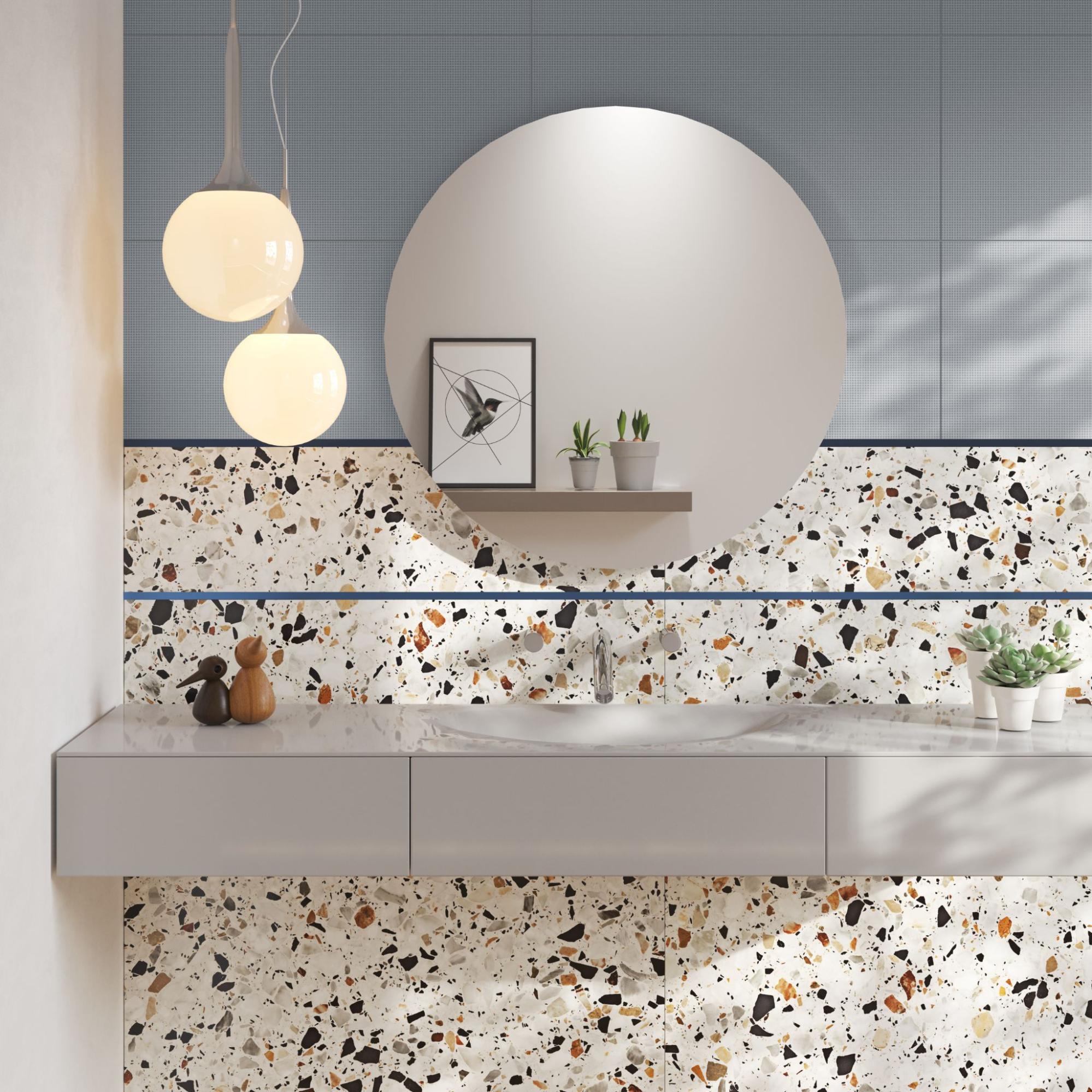 Banheiro moderno com bancada suspensa cinza e espelho redondo. A parede destaca revestimento no estilo granilite em tons claros com detalhes em preto e marrom, combinado com azulejos azul-acinzentados no topo e pendentes luminosos esféricos.
